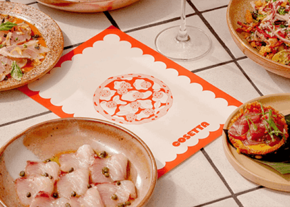 Assorted dishes on a tiled table around a white and red scalloped placemat featuring the Cosetta logo centerpiece.