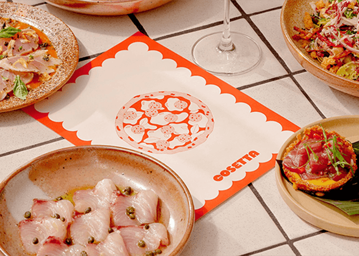 Assorted dishes on a tiled table around a white and red scalloped placemat featuring the Cosetta logo centerpiece.