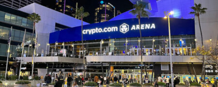 Night view of the crypto.com Arena with a bright blue sign and people outside near planters and palm trees at night.