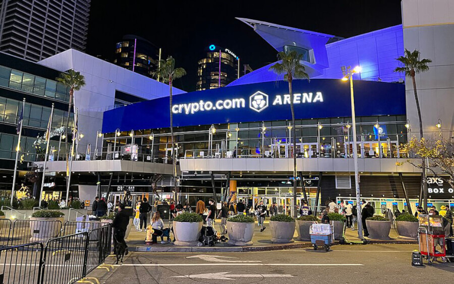 Night view of the crypto.com Arena with a bright blue sign and people outside near planters and palm trees at night.