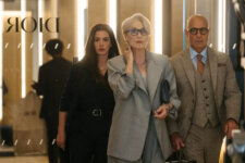 Three stylish professionals walk through a high-end boutique, led by a woman in a light gray suit carrying a handbag while another woman and a man flank her. Dior signage is visible in the background.