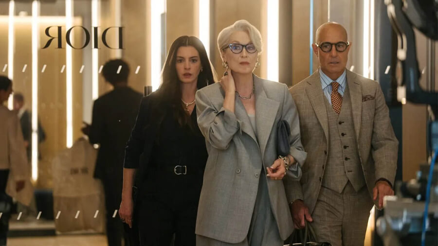 Three stylish professionals walk through a high-end boutique, led by a woman in a light gray suit carrying a handbag while another woman and a man flank her. Dior signage is visible in the background.