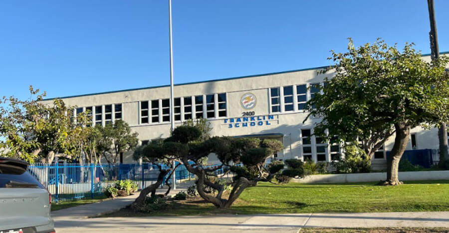 Exterior of Franklin School building with blue fence, trimmed trees, and a parking lot on a sunny day.