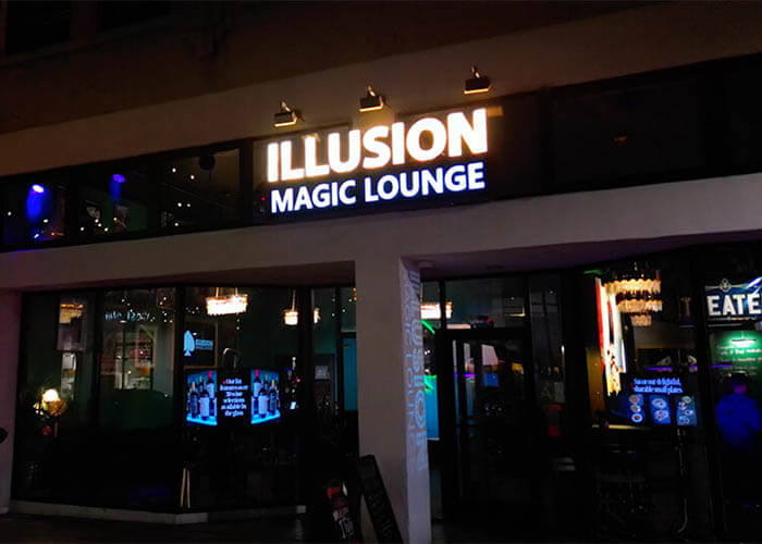Exterior at night of Illusion Magic Lounge with a bright, illuminated sign reading 'ILLUSION MAGIC LOUNGE' above glass doors