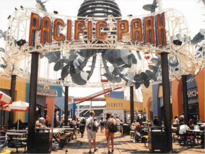 Entrance to Pacific Park with a large orange 'PACIFIC PARK' sign overhead, people walking and seated at picnic tables under a metal arch structure.