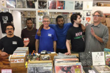 Six men stand behind a cluttered vinyl record shop counter, with shelves of records above them and boxes of records in front. One man on the left holds a small red box while others pose for the photo.