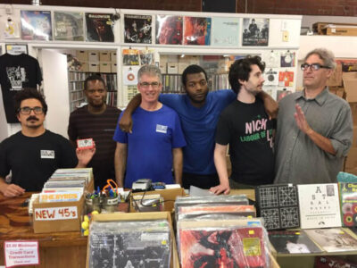 Six men stand behind a cluttered vinyl record shop counter, with shelves of records above them and boxes of records in front. One man on the left holds a small red box while others pose for the photo.