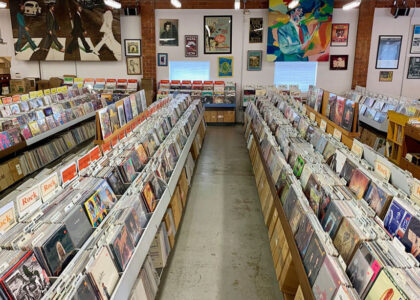 Rows of vinyl records in a record store with rock-label sections and wall posters above the aisles.