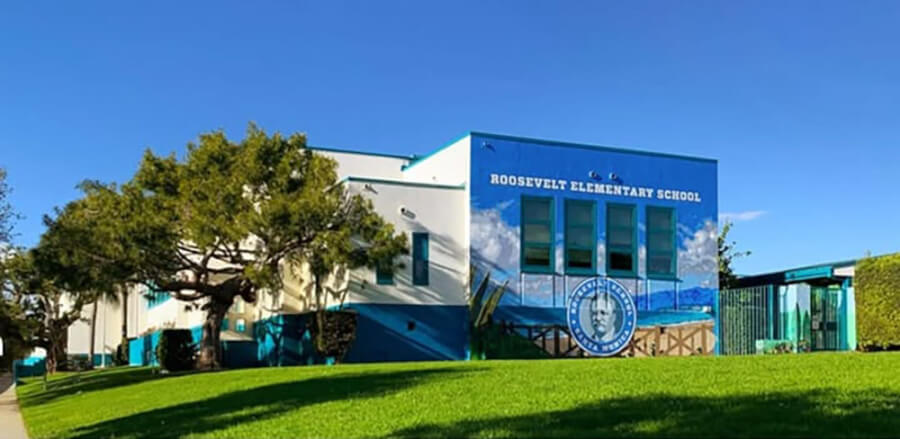 Exterior view of Roosevelt Elementary School with a blue mural on the façade, green lawn, and trees under a clear sky.