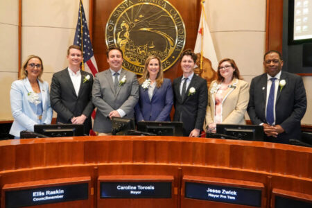 Seven officials in suits stand behind a curved wooden dais in a council chamber with a large city seal behind them.