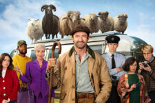 Group of diverse adults pose in front of a retro bus with a flock of sheep perched on the roof; a man center-left holds a wooden crook, smiling. Group photo from a quirky rural scene featuring several characters and a bus topped with sheep. A varied cast stands in front of a silver bus topped with sheep, led by a man in a brown coat holding a shepherd’s staff. People from different backgrounds stand before a vintage bus with sheep on top, suggesting a humorous, rural adventure. Front-facing ensemble shot with a bus carrying multiple sheep on its roof; central man in tan coat smiles at the camera.