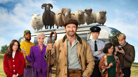Group of diverse adults pose in front of a retro bus with a flock of sheep perched on the roof; a man center-left holds a wooden crook, smiling. Group photo from a quirky rural scene featuring several characters and a bus topped with sheep. A varied cast stands in front of a silver bus topped with sheep, led by a man in a brown coat holding a shepherd’s staff. People from different backgrounds stand before a vintage bus with sheep on top, suggesting a humorous, rural adventure. Front-facing ensemble shot with a bus carrying multiple sheep on its roof; central man in tan coat smiles at the camera.