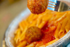 Vitos pasta and meatballs | Santa Monica Sun Meatball on a fork above penne pasta with tomato sauce in a foil takeout container, close-up view.