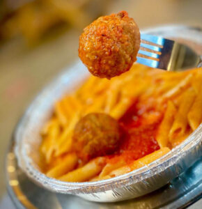Meatball on a fork above penne pasta with tomato sauce in a foil takeout container, close-up view.