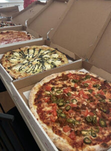 Several large pizzas sit in open cardboard boxes on a table, with the front pizza topped with jalapeños, tomatoes, and sausage/Ground meat blend highlight visible at the edges.