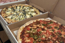 Vitos specialty pizzas | Santa Monica Sun Several large pizzas sit in open cardboard boxes on a table, with the front pizza topped with jalapeños, tomatoes, and sausage/Ground meat blend highlight visible at the edges.