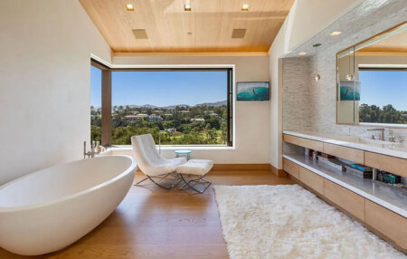 859 Woodacres Road bathroom | Santa Monica Sun Bright, modern bathroom with a freestanding oval tub, white lounge chair, and a large window showing a green landscape.