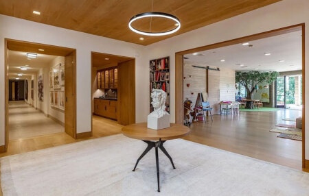 859 Woodacres Road entryway | Santa Monica Sun Modern lobby with a round wooden table and a white bust sculpture, open doorways to a kitchen on the left and a play area with a tree on the right.