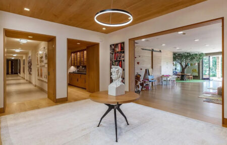 Modern lobby with a round wooden table and a white bust sculpture, open doorways to a kitchen on the left and a play area with a tree on the right.