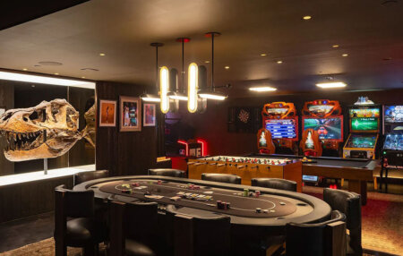 859 Woodacres Road game room | Santa Monica Sun Dimly lit entertainment lounge with a circular poker table, leather chairs, and a wall-mounted dinosaur skeleton exhibit behind.