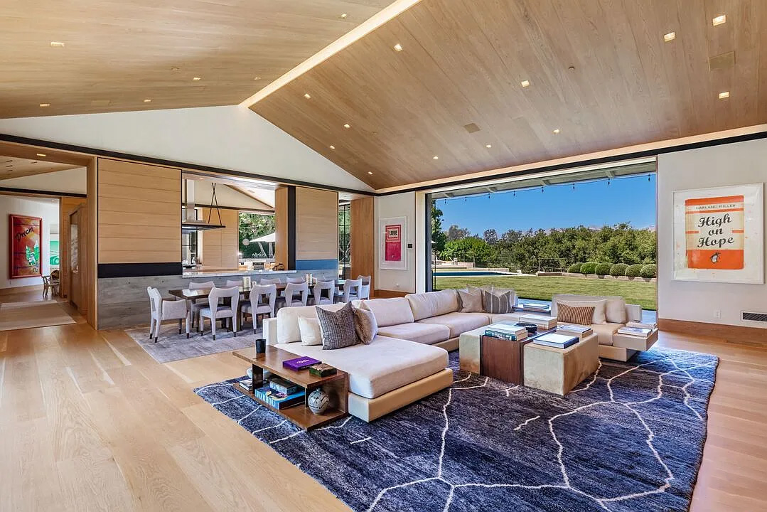 859 Woodacres Road living room | Santa Monica Sun Open-concept living room with a beige sectional, coffee tables, and a blue rug, framed by a large glass wall overlooking a green yard.