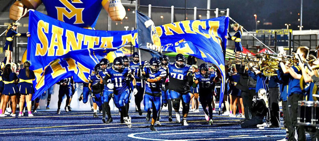 High school football players sprint onto the field under large blue and yellow banners held by cheerleaders and fans on a blue turf field.