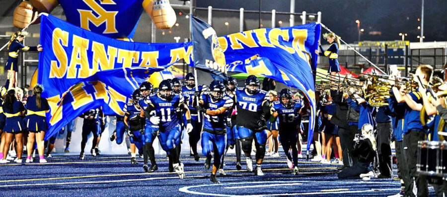High school football players sprint onto the field under large blue and yellow banners held by cheerleaders and fans on a blue turf field.