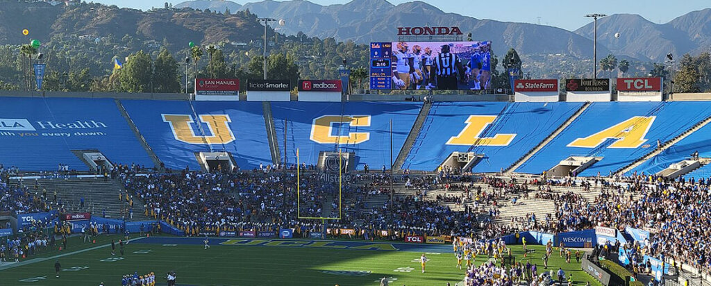 Wide view of a football stadium with blue seats spelling UCLA, a large crowd, and players on the field under a big video board.