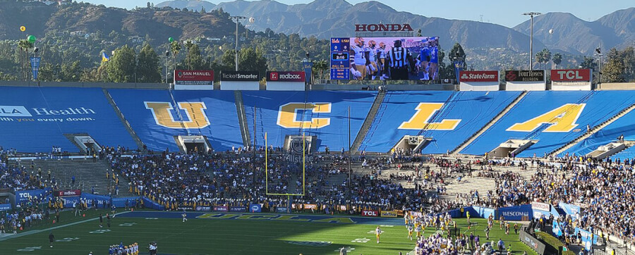 Wide view of a football stadium with blue seats spelling UCLA, a large crowd, and players on the field under a big video board.