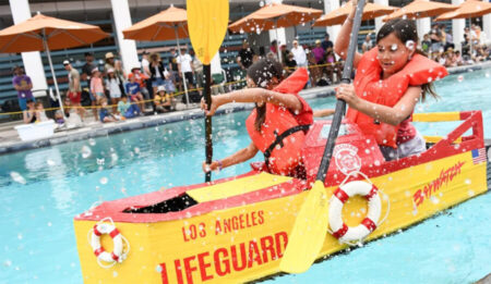 Two children in bright orange life jackets paddle a yellow lifeguard boat in a pool while spectators watch from the edge.