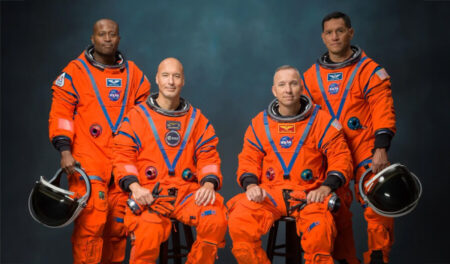 Four astronauts in orange spacesuits pose for a NASA team photo against a dark backdrop, with helmets nearby at the sides of the frame.