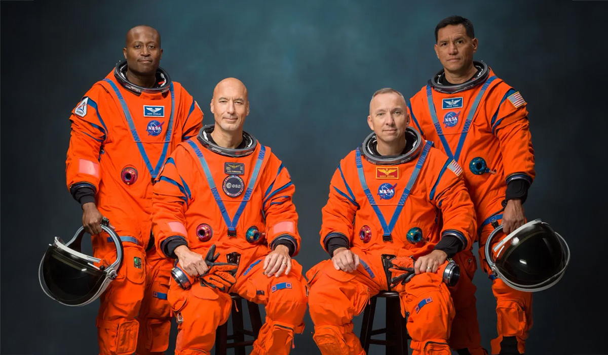 artemis-crew-randy-bresnik | Santa Monica Sun Four astronauts in orange spacesuits pose for a NASA team photo against a dark backdrop, with helmets nearby at the sides of the frame.