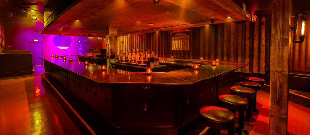 Dimly lit nightclub bar with a glossy U-shaped counter, row of stools, and colored ambient lighting behind it.