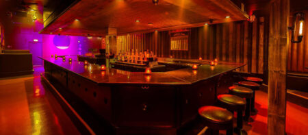 Dimly lit nightclub bar with a glossy U-shaped counter, row of stools, and colored ambient lighting behind it.