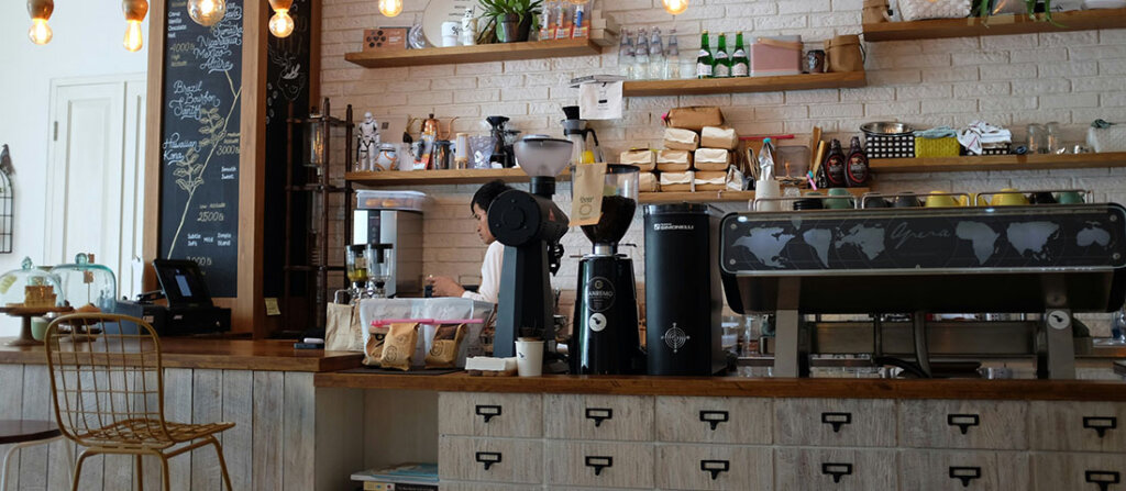 Cafe counter with barista at an espresso machine, shelves of coffee bags and gear against a white brick wall.