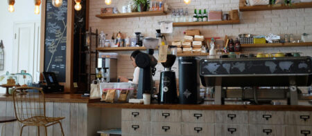 Cafe counter with barista at an espresso machine, shelves of coffee bags and gear against a white brick wall.