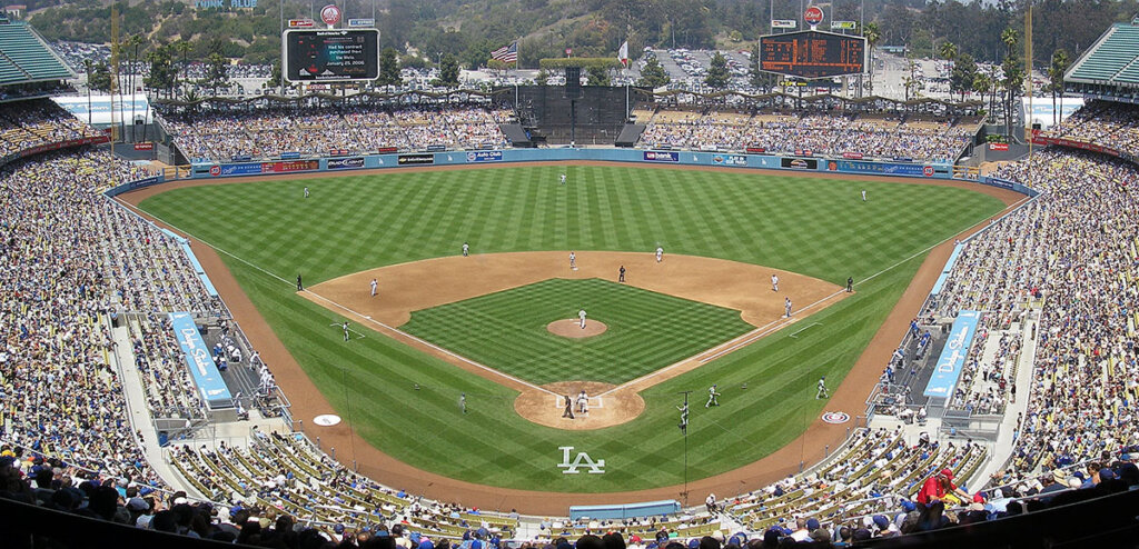 Panoramic view of a baseball game at Dodger Stadium, with a full crowd, players positioned on the diamond, and the LA logo near home plate.