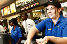 McDonald's cashier in blue uniform taking cash from a customer at the counter, with menu boards behind.