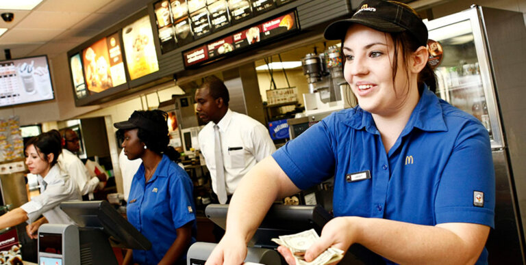 McDonald's cashier in blue uniform taking cash from a customer at the counter, with menu boards behind.