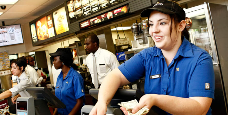 McDonald's cashier in blue uniform taking cash from a customer at the counter, with menu boards behind.