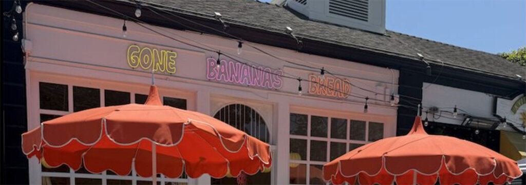 Front of a bakery storefront with pink neon sign reading 'Gone Bananas Bread' and two orange patio umbrellas outside.