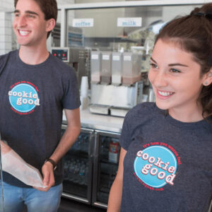Two smiling staff members in matching gray t-shirts with a Cookie Good logo, standing behind a cafe counter with coffee and milk machines in the background.