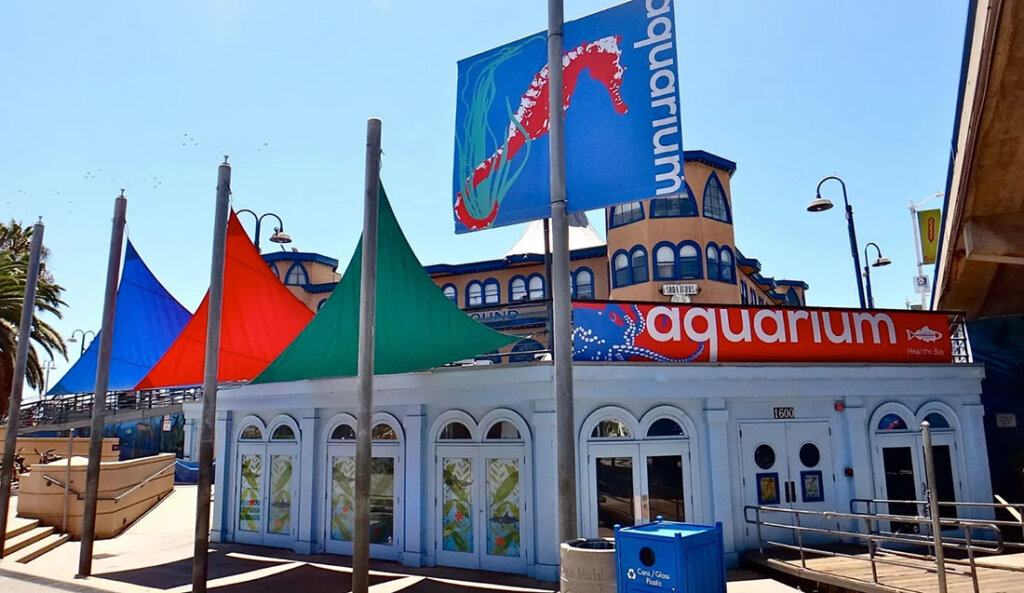 Outdoor aquarium building with white arches and a bright red sign reading 'aquarium' under multicolored fabric sails (blue, red, green).