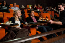 A couple sits in orange theater seats smiling as a waiter presents a tray with three burgers; other patrons relax in the background at a cinema bar area.