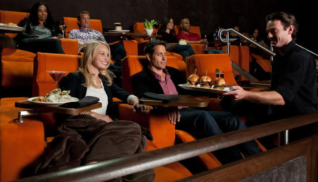 A couple sits in orange theater seats smiling as a waiter presents a tray with three burgers; other patrons relax in the background at a cinema bar area.