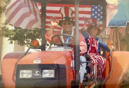 Two people ride an orange tractor in a patriotic parade, waving at onlookers with American flags in the background.