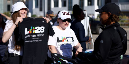 Volunteers at an outdoor event distribute T-shirts, one woman proudly holding a black shirt for a Delta Airlines volunteer program.