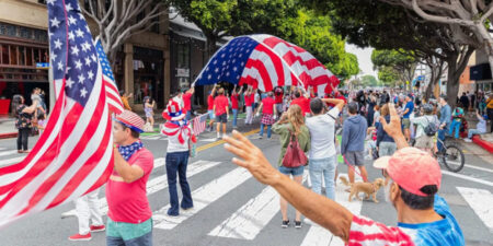 People in a street parade hold up a large American flag, amid a crowd on a crosswalk and storefronts in the background.