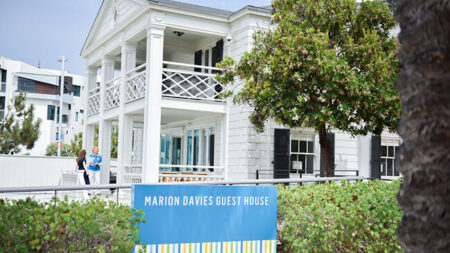 White two-story guest house with wraparound porch and a blue sign reading 'Marion Davies Guest House' in front of greenery.