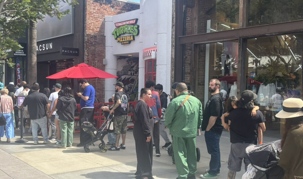 People line up outside a storefront with a Teenage Mutant Ninja Turtles Pizzeria sign and a red umbrella on a city street.